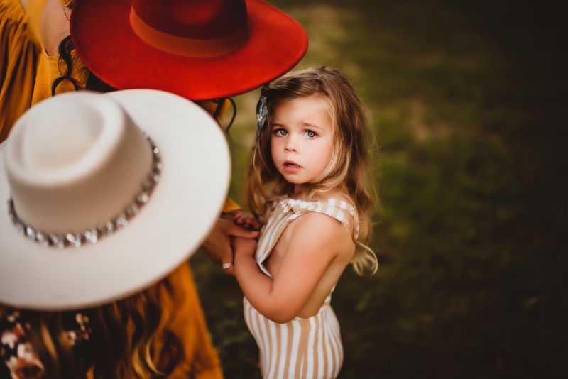 Family Photographer, a little girl stands in the grass with two other women wearing hats